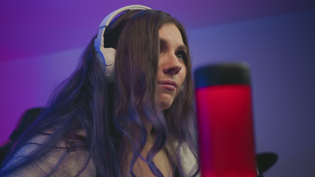 closeup of tense lady wearing white headphones focused intensely while playing video game in moody lighting with red condenser microphone glowing beside her in modern streaming setup