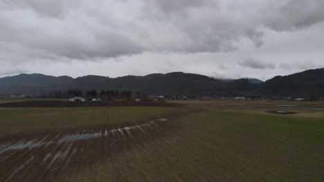 Farmland-fields-of-crops-rows-of-vegetables-on-farm-in-country-valley-surrounded-by-mountains-on-cloudy-day-Aerial-ascending-revealing-farm-mountains-in-background