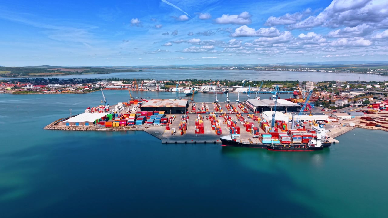 Burgas, Bulgaria, 1 May 2026: Container Ship Docked At Industrial Seaport Terminal. Aerial view of container ship docked beside stacked containers, cranes, warehouses, and blue harbor water.