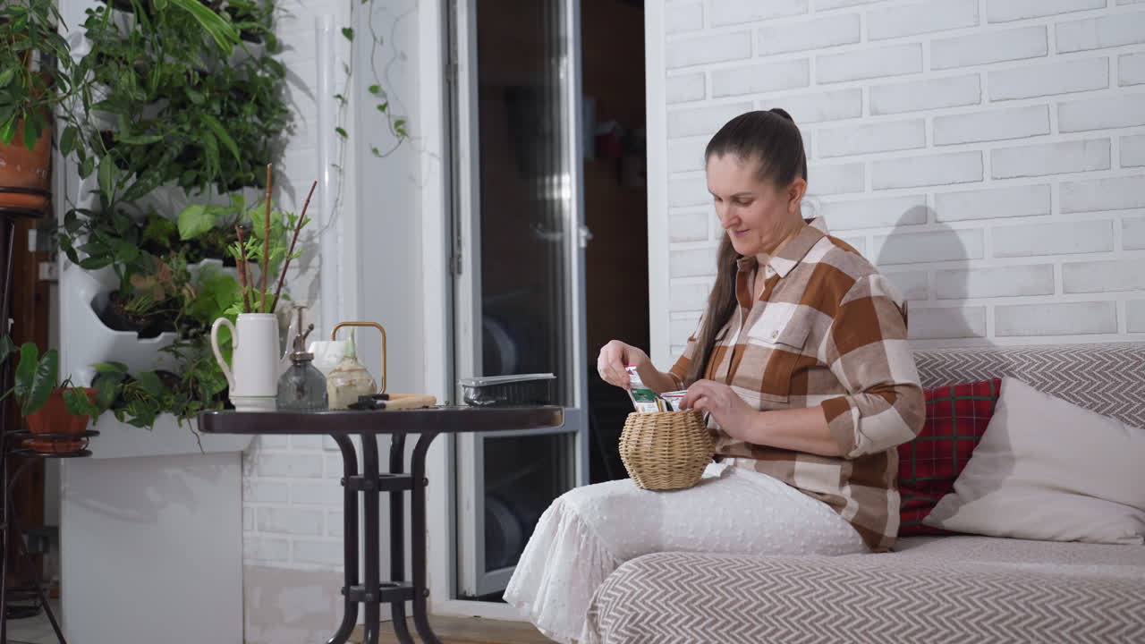 Floral caretaker seated on sofa near round table arranging plant seed packet inside small basket on lap against white brick wall background with lush indoor greenery, sorting botanical tags