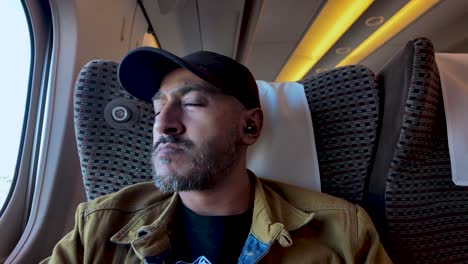 hombre del sur de asia con auriculares y gorra de béisbol mirando pensativo por la ventana del tren mientras viaja