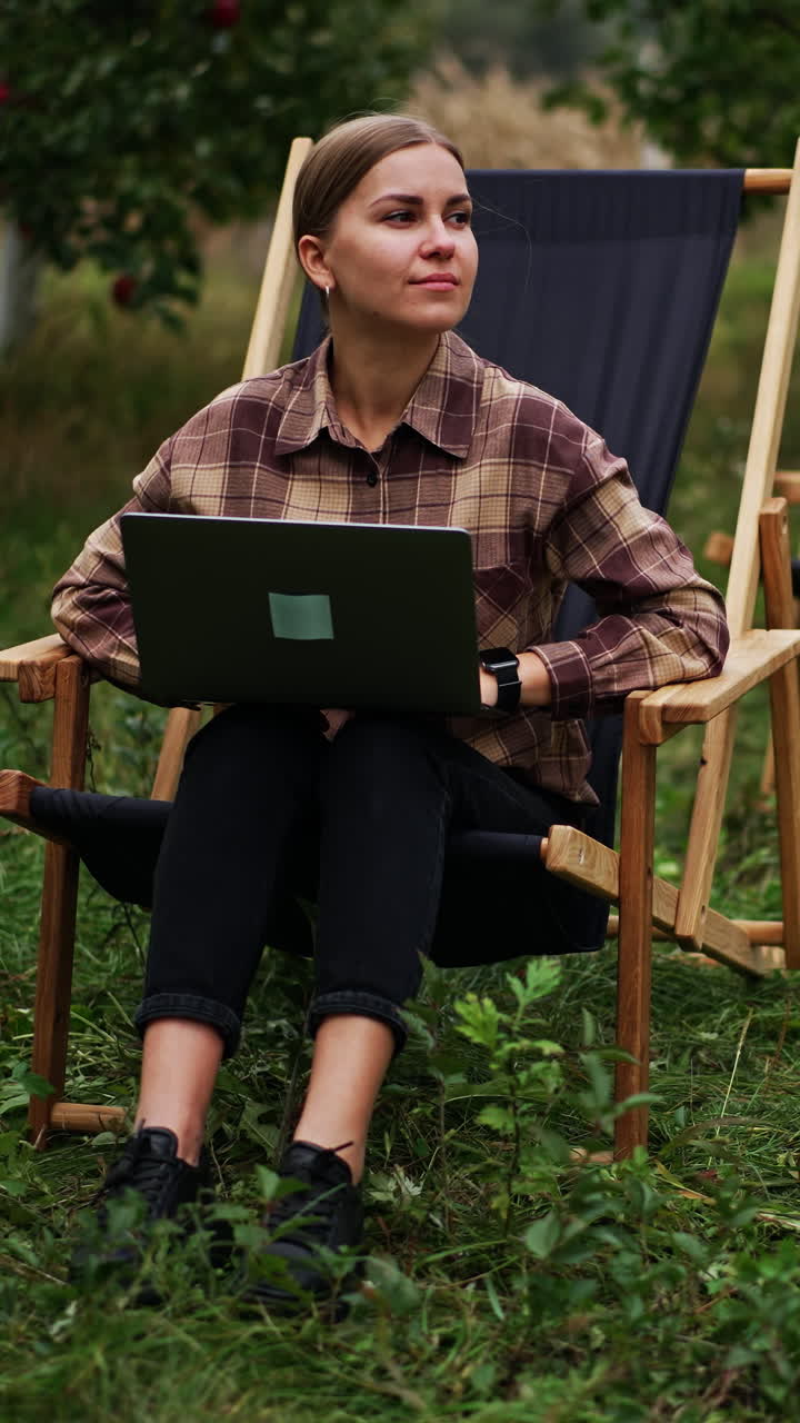 Pensive lady in plaid shirt sitting in garden chair working on laptop. Freelance working woman thinking over some work issues. Nature backdrop. Vertical video
