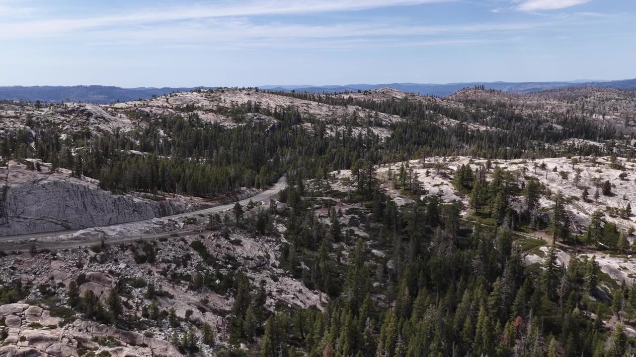 Plan drone en approche au-dessus de montagnes de granit accidentées, de routes sinueuses et d'une forêt dense de conifères près de Silver Lake Pioneer, Californie, capturé sous une lumière du jour éclatante avec des vues paysagères étendues.
