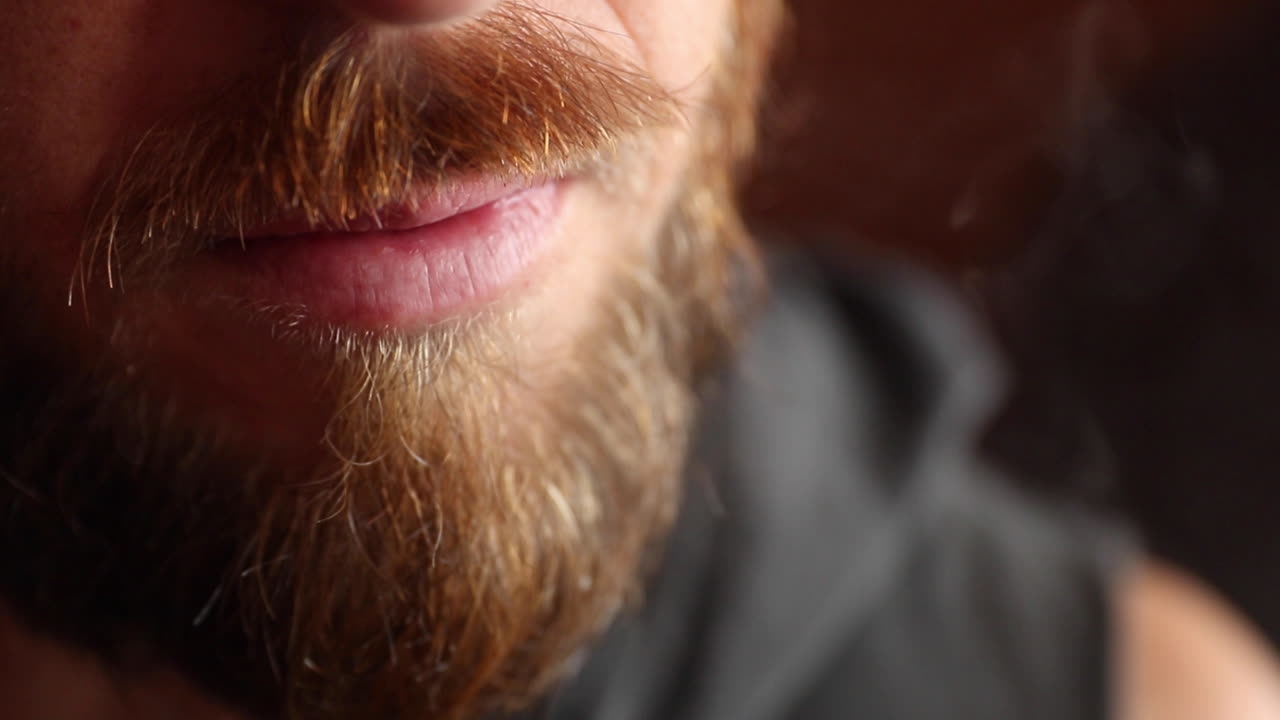 primer plano de un hombre barbudo que lleva un chaleco negro y fuma un cigarrillo enrollado