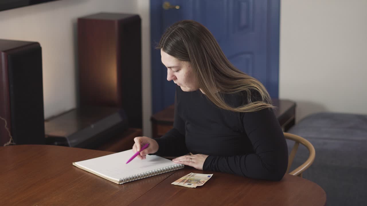 Donna serena esamina un piano di bilancio a casa, scrivendo su un quaderno accanto a contanti e un computer per la finanza personale, i risparmi o la pianificazione domestica.