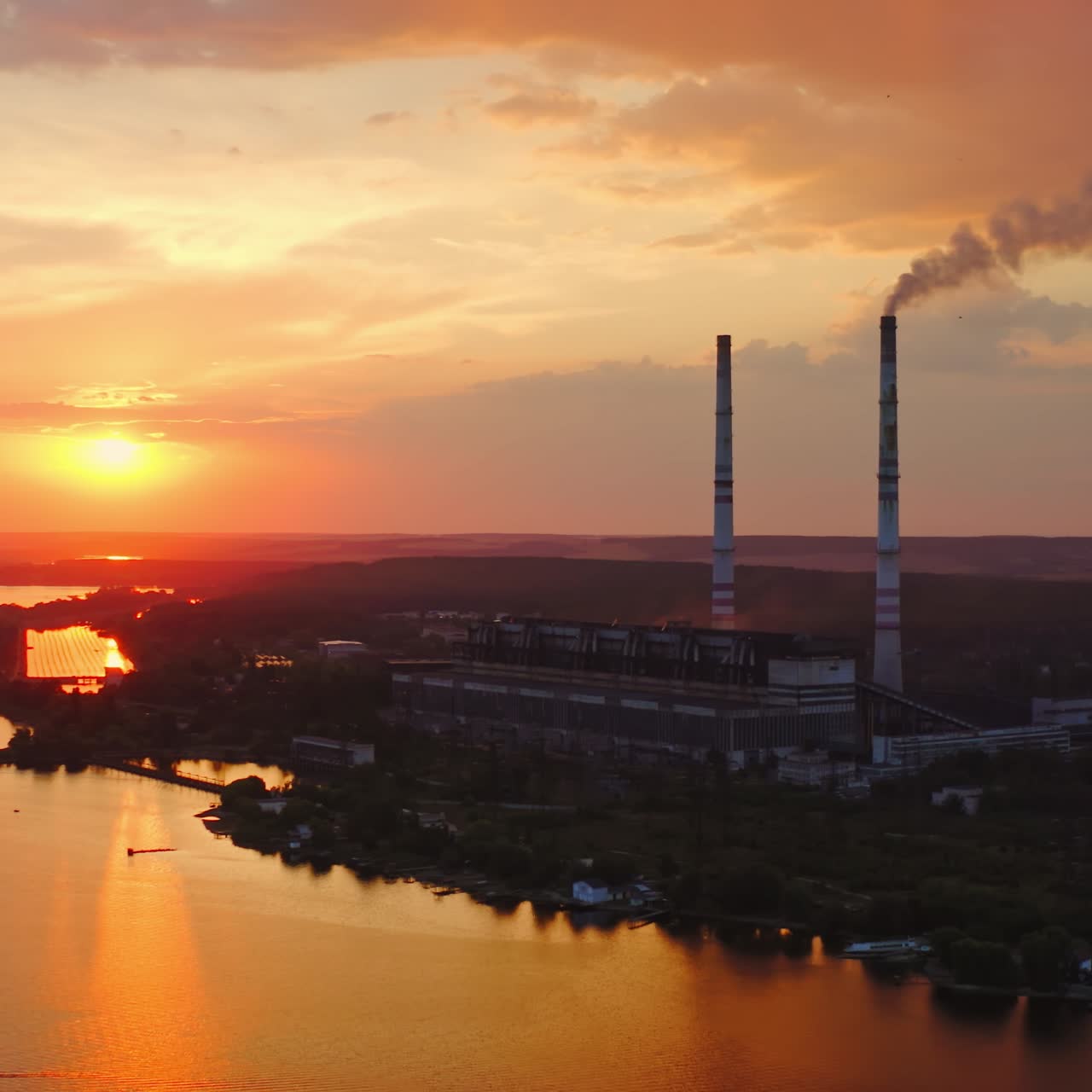Factory near the river at sunset. Industrial plant with smoking pipe against the evening sky. Ecological problems. Motion camera back. Aerial view.