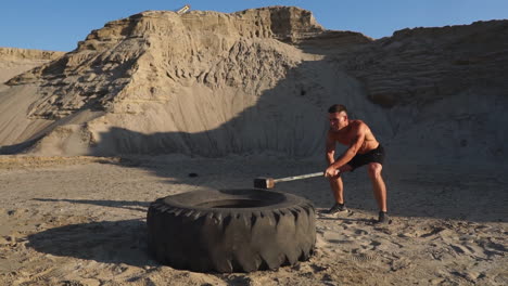 atleta muscular hombre fuerte golpea un martillo en una rueda enorme en las montañas arenosas en cámara lenta. entrenamiento de fuerza y resistencia para luchadores