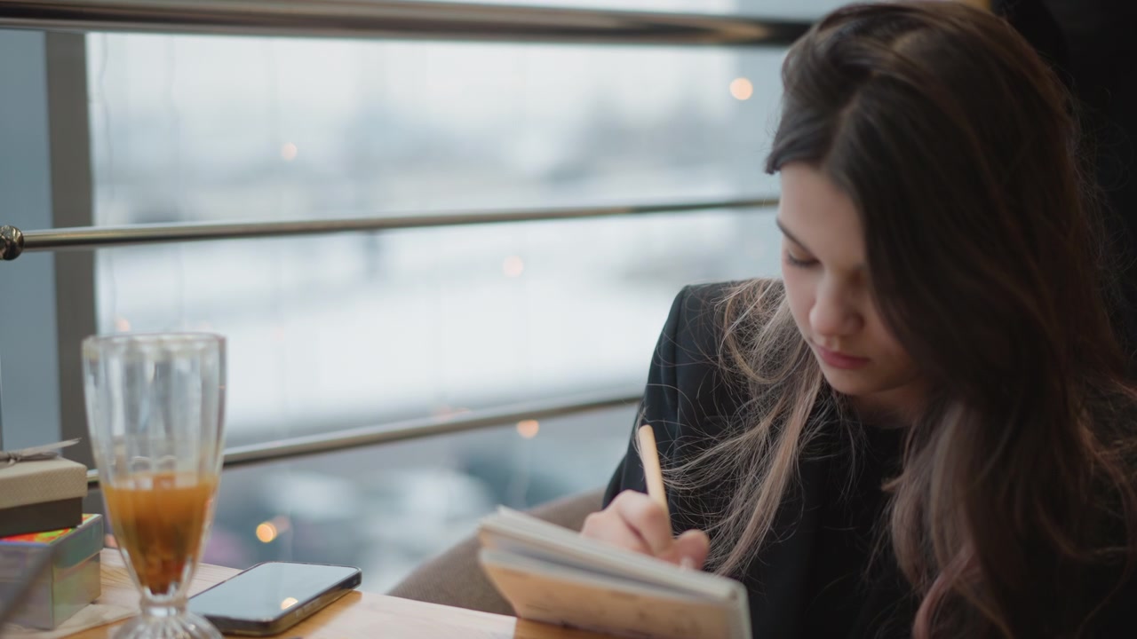 Primo piano medio mostra una donna seduta a un tavolo di legno di un caffè, che scrive su un quaderno accanto a una finestra luminosa; il movimento della penna, lo sguardo abbassato e la luce interna soffusa suggeriscono una tranquilla presa di appunti e una pianificazione concentrata.