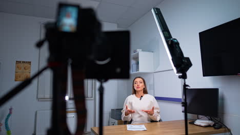 Confident good-looking female speaker sits at desk talking. Presenter speaks to camera recording video content.