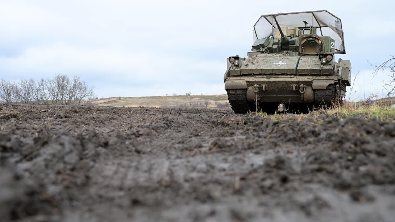 A low-angle shot of a US-made M2 Bradley on muddy terrain in Ukraine. It's adapted with a medical cross for CASEVAC and cage armor for defense against enemy drones on a muddy terrain