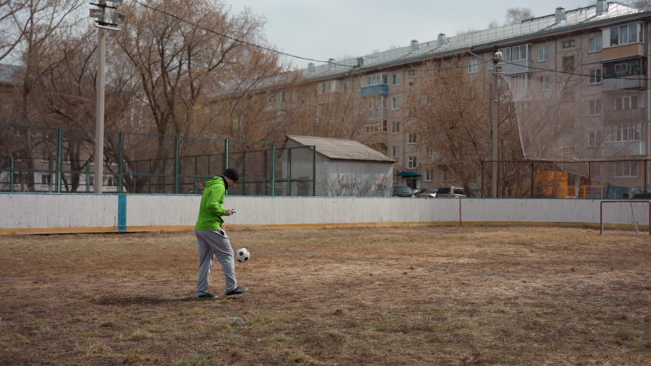 Молодой белый мужчина тренируется в дриблинге на открытом поле, в зеленой куртке и шапке; осенняя трава, городские многоквартирные дома на заднем плане, индивидуальная тренировка, отработка техники владения мячом в холодную погоду