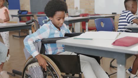 Diverse-male-teacher-and-happy-schoolboy-in-wheelchair-writing-in-classroom-at-elementary-school