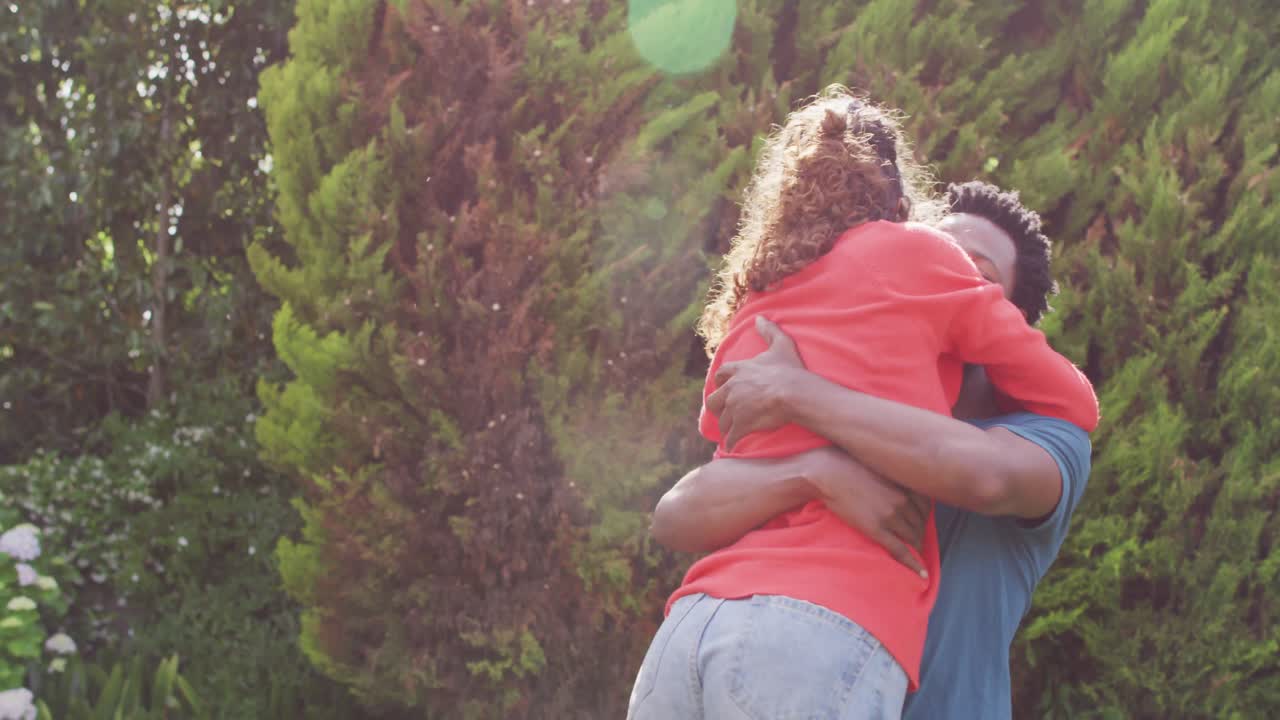 Happy biracial man hugging his fiance with engagement ring on hand in garden in sun