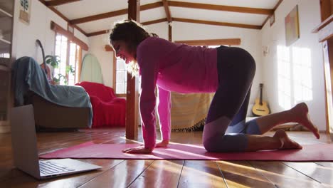 Relaxed-mixed-race-woman-practicing-yoga,-kneeling-and-stretching-in-sunny-cottage-bedroom