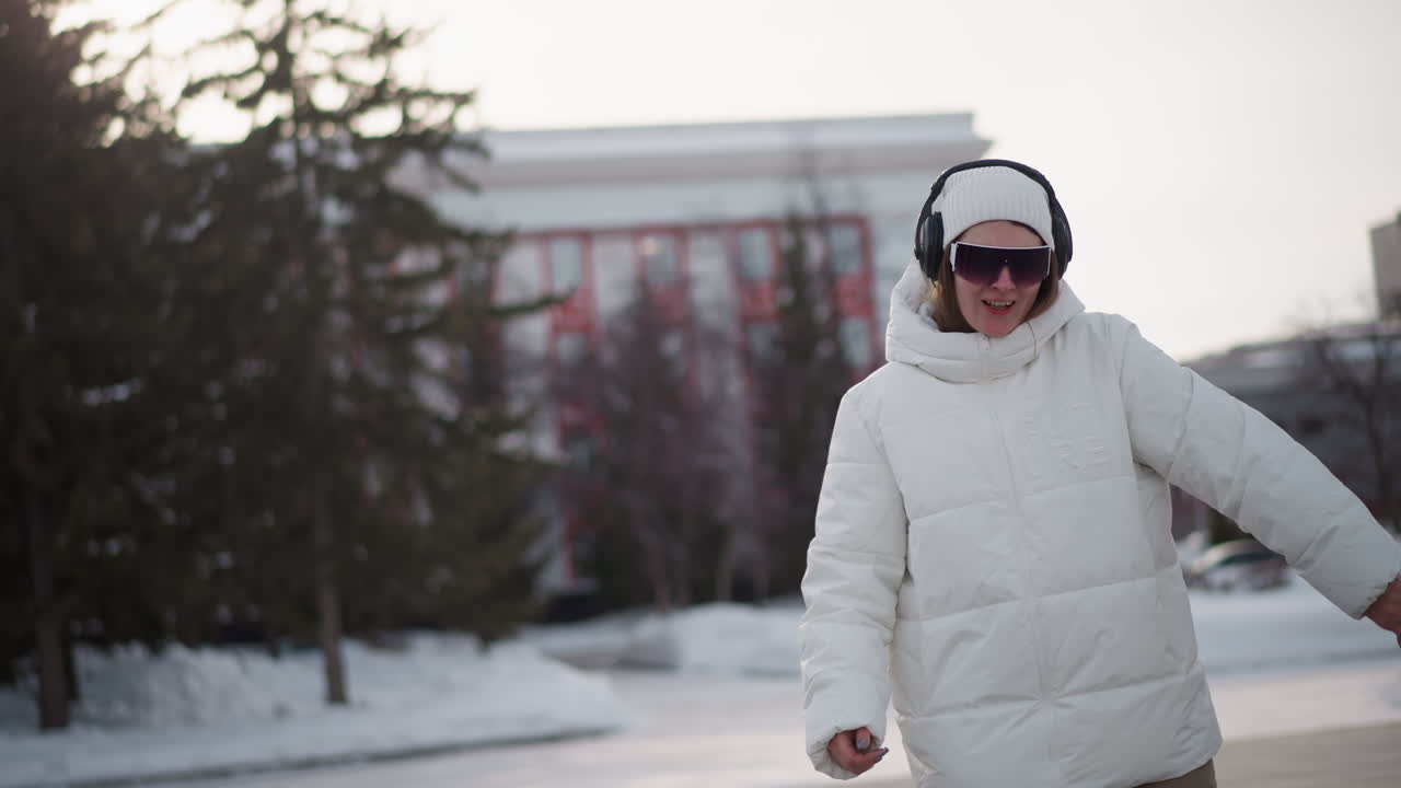 Closeup of school girl wearing fluffy white coat, beanie, sunglasses and headphones, dancing around with arms extended, joyful expression with playful movement on snow dusted urban plaza