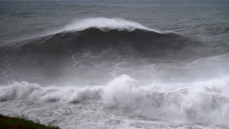 Huge-big-wave-breaking-in-Nazaré,-view-from-the-beach-with-spray-in-the-foreground