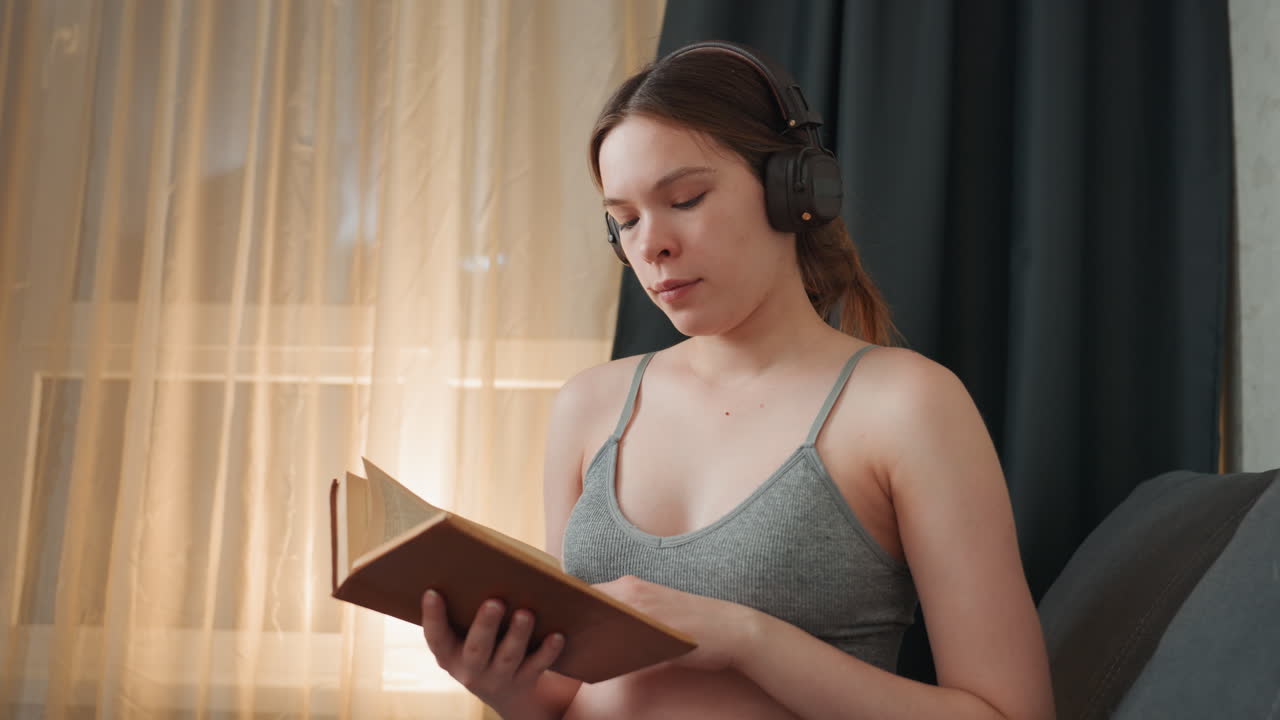 Pregnant caucasian woman reading with headphones, closeup portrait, knit top, focused expression, holding book while listening to audiobook, soft golden light through curtains, quiet prenatal