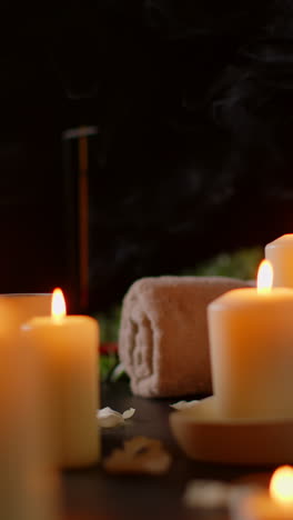 Vertical-Video-Still-Life-Of-Lit-Candles-With-Scattered-Petals-Incense-Stick-And-Soft-Towels-Against-Dark-Background-As-Part-Of-Relaxing-Spa-Day-Decor