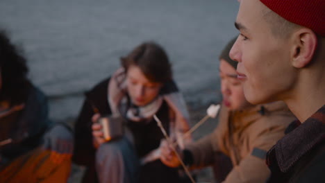 Rear-View-Of-A-Teenage-Boy-Roasting-And-Eating-Marshmallows-Sitting-Around-Bonfire-With-Her-Friends