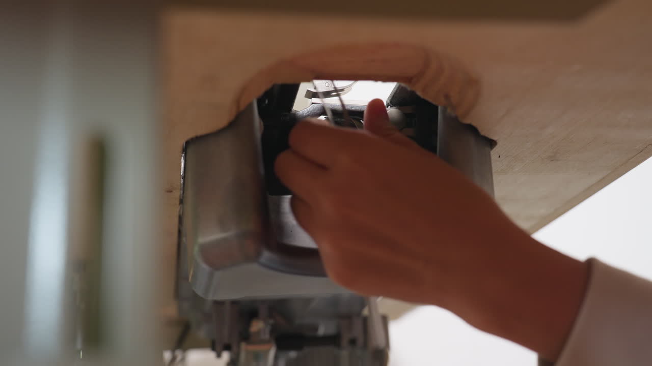 Close view of seamstress hand pulling thread from beneath weaving machine while performing maintenance, focusing on intricate under table angle and technical adjustment in sewing workshop environment