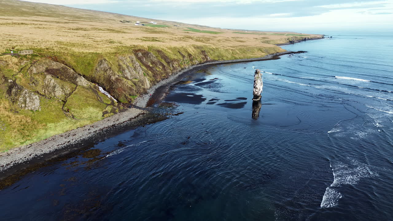 Dronefoto av Hvítserkur, den berømte basalte havstakken som stiger opp fra havet langs Vatnsnes-halvøya på Nord-Island. Bølger ruller forsiktig inn over den svarte sandstranden, mens klipper og gresskledde kystsletter strekker seg langs strandlinjen.