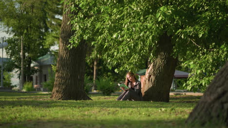 woman sitting outdoors, leaning against tree on grassy field, reading book under warm sunlight, tree leaves sway gently in breeze, background shows residential building and lamp post