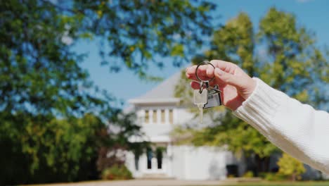 a hand with a keychain in the shape of a small house and keys. against the background of a typical house in the american suburban style