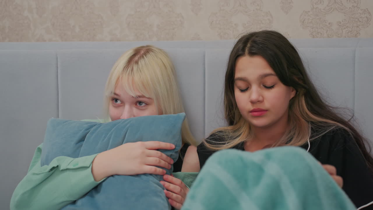 young girls sitting on bed watching horror movie looking visibly frightened, brunette clutching blanket nervously while blonde girl hides behind pillow
