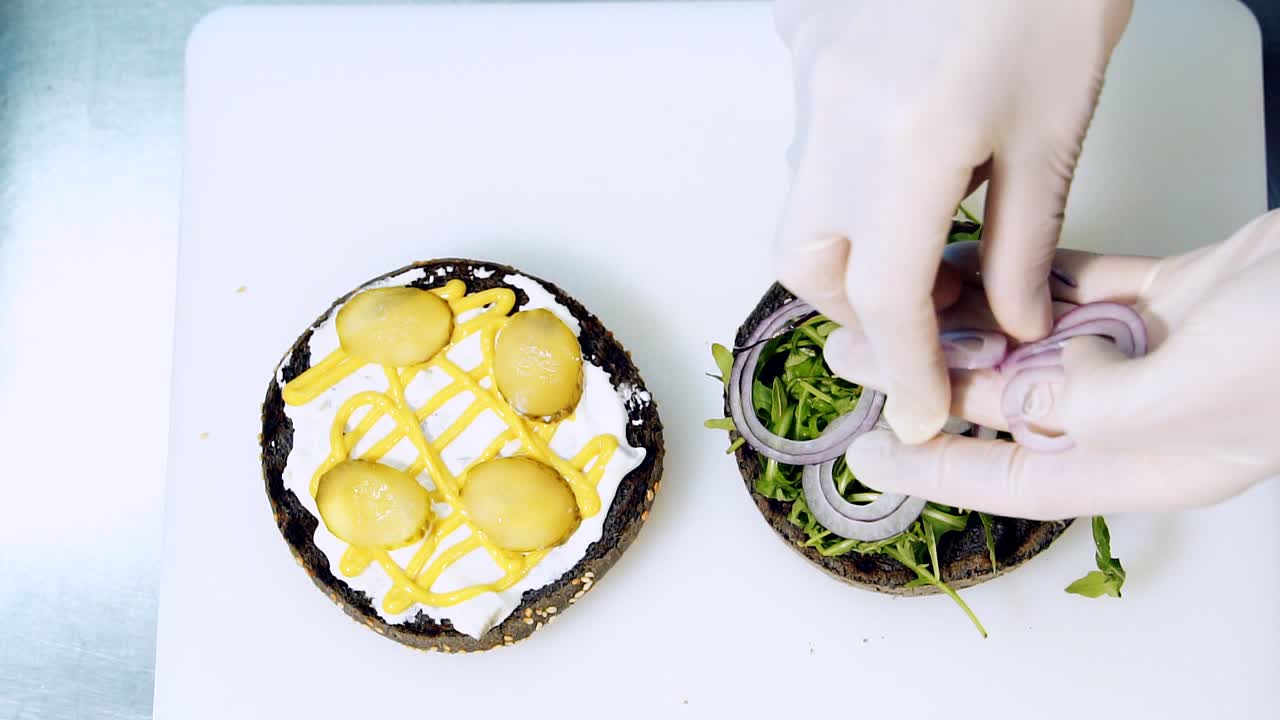 Culinary procedure of making tasty black burger. Chef puts onion rings while making vegan hamburger. Cooking healthy food. Top view