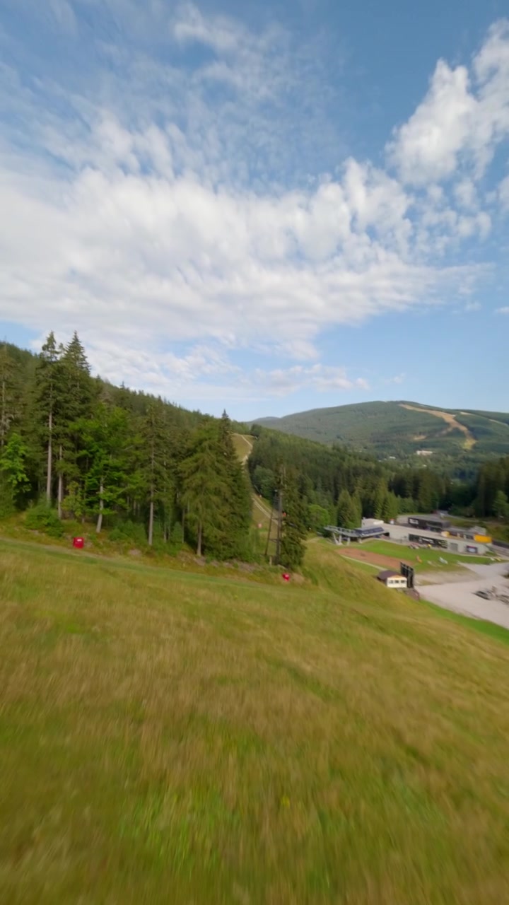 Ripresa dinamica verticale con drone FPV che si libra su lussureggianti foreste di pini e piste da sci erbose nelle montagne ceche durante l'estate. Movimento ad alta energia su un resort di montagna