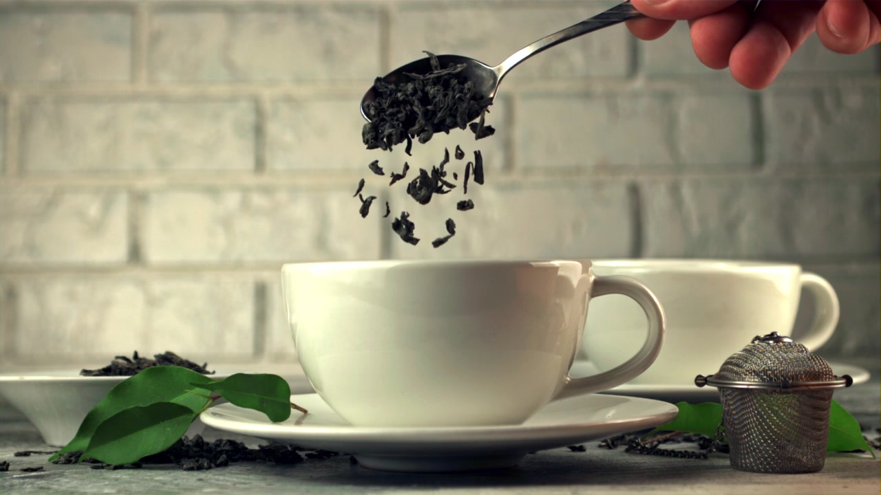 Foglie di tè secche che cadono da un cucchiaio in una tazza da tè bianca in slow motion