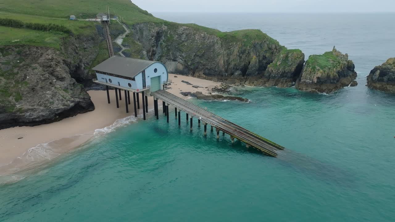 RNLI lifeboat station with long slipway into clear blue sea next to clean sandy beach with seagulls flying. Mother Ivey's Bay, Cornwall, UK.