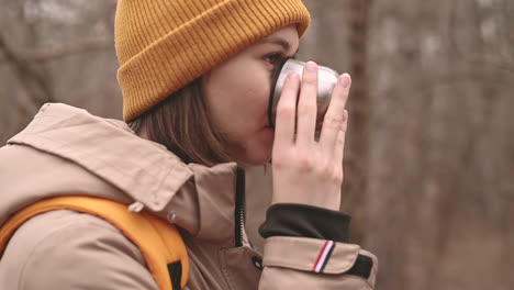 a young girl in a yellow woolen hat drinks a hot beverage in the forest