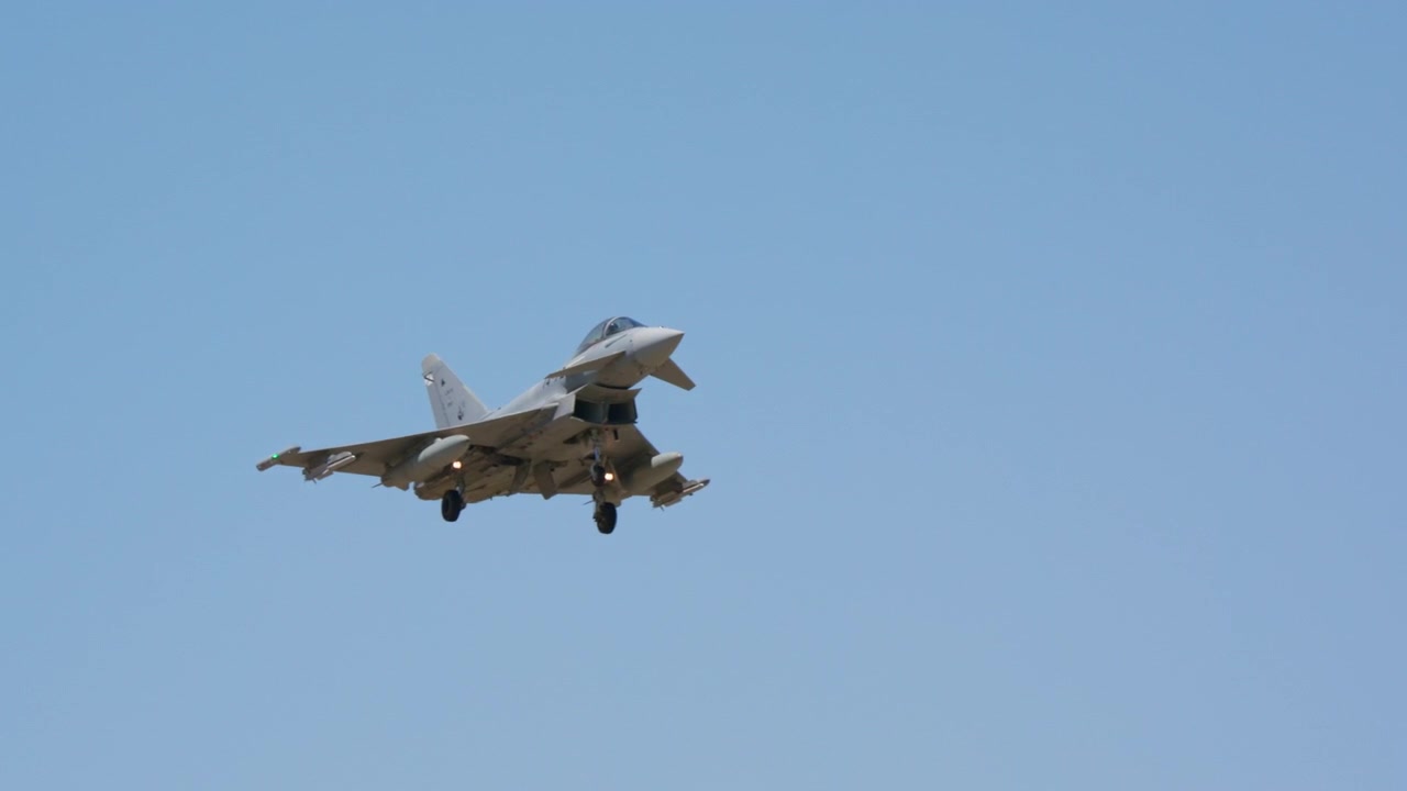 Jet tempur militer mendekat dalam gerakan lambat dengan roda pendaratan diturunkan, terbang menuju kamera dengan latar belakang langit biru cerah