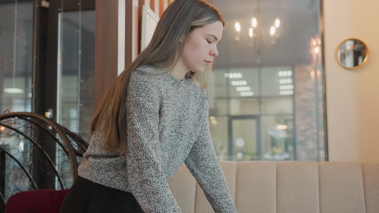 Close up of pretty lady in restaurant placing beverage on table and sitting comfortably on chair with mirror reflection of woman passing by behind her, creating calm indoor ambiance