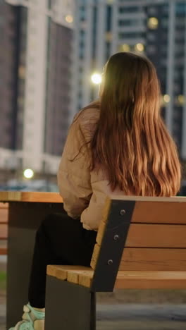 fotografía trasera de una chica con el cabello largo y marrón en una chaqueta de melocotón y pantalones negros, sentada en un banco del parque. ella está levantando la cabeza ligeramente, con edificios altos y borrosos y personas distantes en el fondo