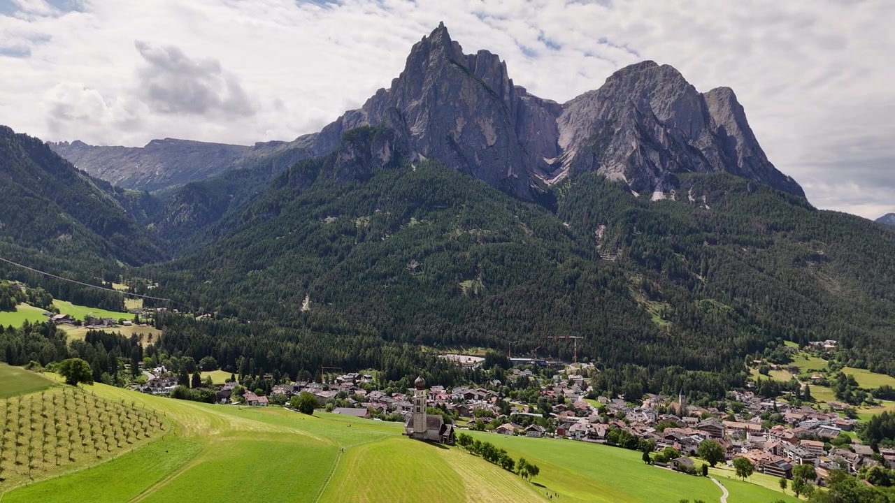 ドロミテ山脈の壮大な峰々に囲まれた、魅力的な歴史あるカステルロットの町を4Kドローンで空撮。象徴的な聖ヴァレンティン教会の鐘楼が印象的です。