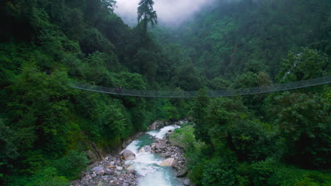Male-tourist-crosses-suspension-bridge-Nepal,-waterfall,-white-river,-forest,-landscapes,-greenery,-monsoon-season,-rural-village-relaxing-scenery-beauty-4K