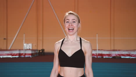 excited blonde sportswoman jumping and cheering to celebrate victory while looking at camera in an indoor sport facility