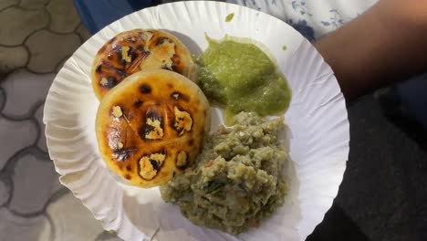 video depicts a man holding a plate of savory litti chokha smeared with ghee and served with roasted mashed eggplant or baingan bharta and a green spicy chutney