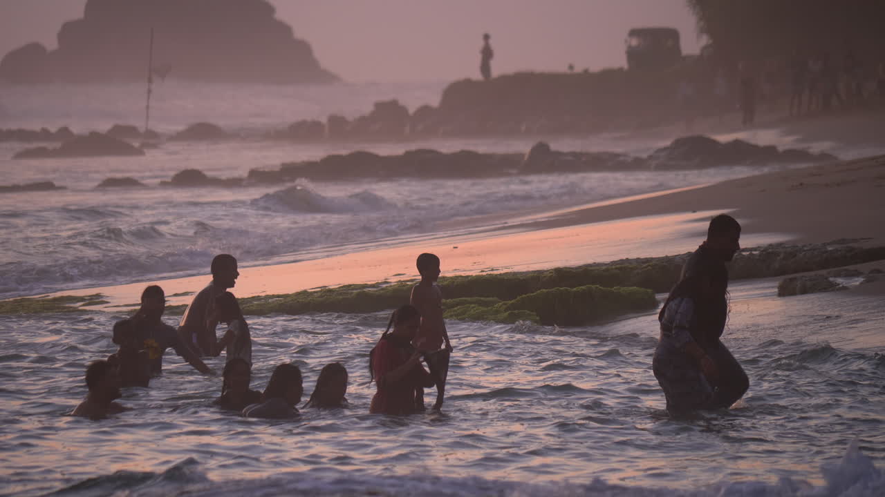 People bathe in the ocean at sunset during the golden hour. The sky is painted pink. Waves crash on the shore