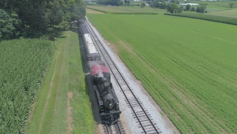 vista aérea de un motor de vapor de 1910 con un tren de pasajeros que echa humo viajando a lo largo de la campiña amish en un día soleado de verano visto por un dron