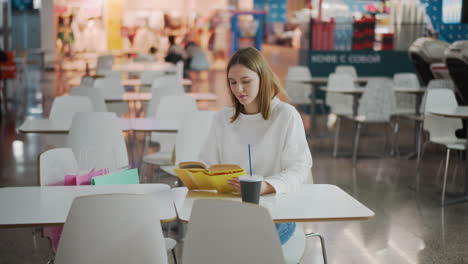 young woman seated at table reading book in vibrant mall surrounded by shopping bags, soft lighting, and colorful background with other shoppers visible in distant seating