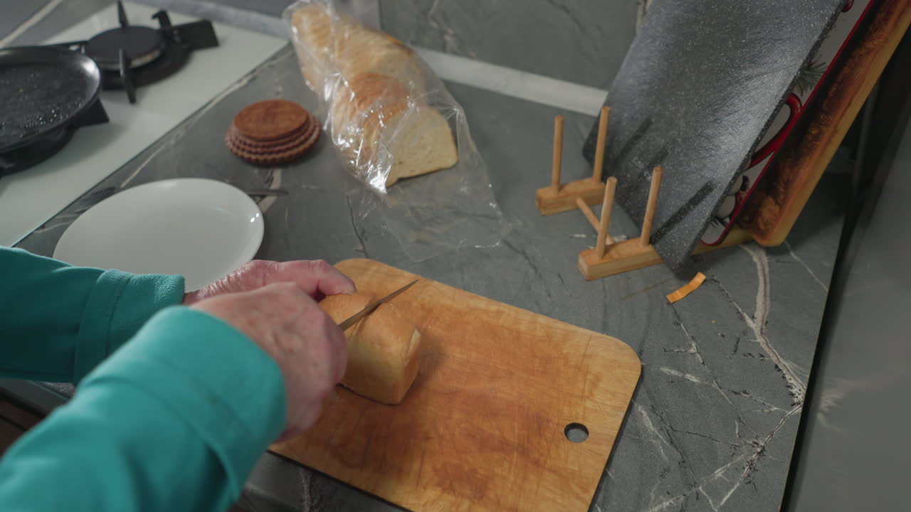 Older woman slicing fresh bread on wooden cutting board with a knife, kitchen counter and kitchen tools visible, preparing food at home with modern appliances and cookware