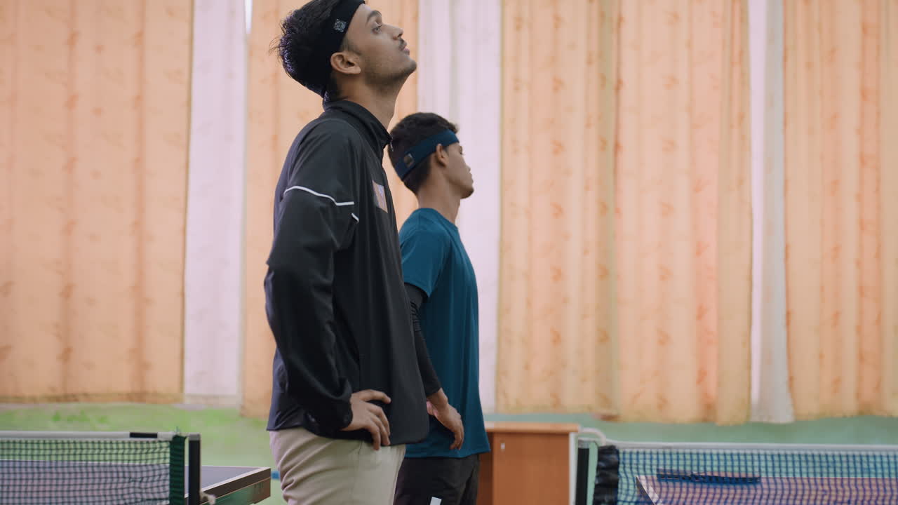 Side view of two friends in sportswear performing neck flexibility exercise near tennis table, warming up muscles with concentration and discipline before practice inside indoor sports hall