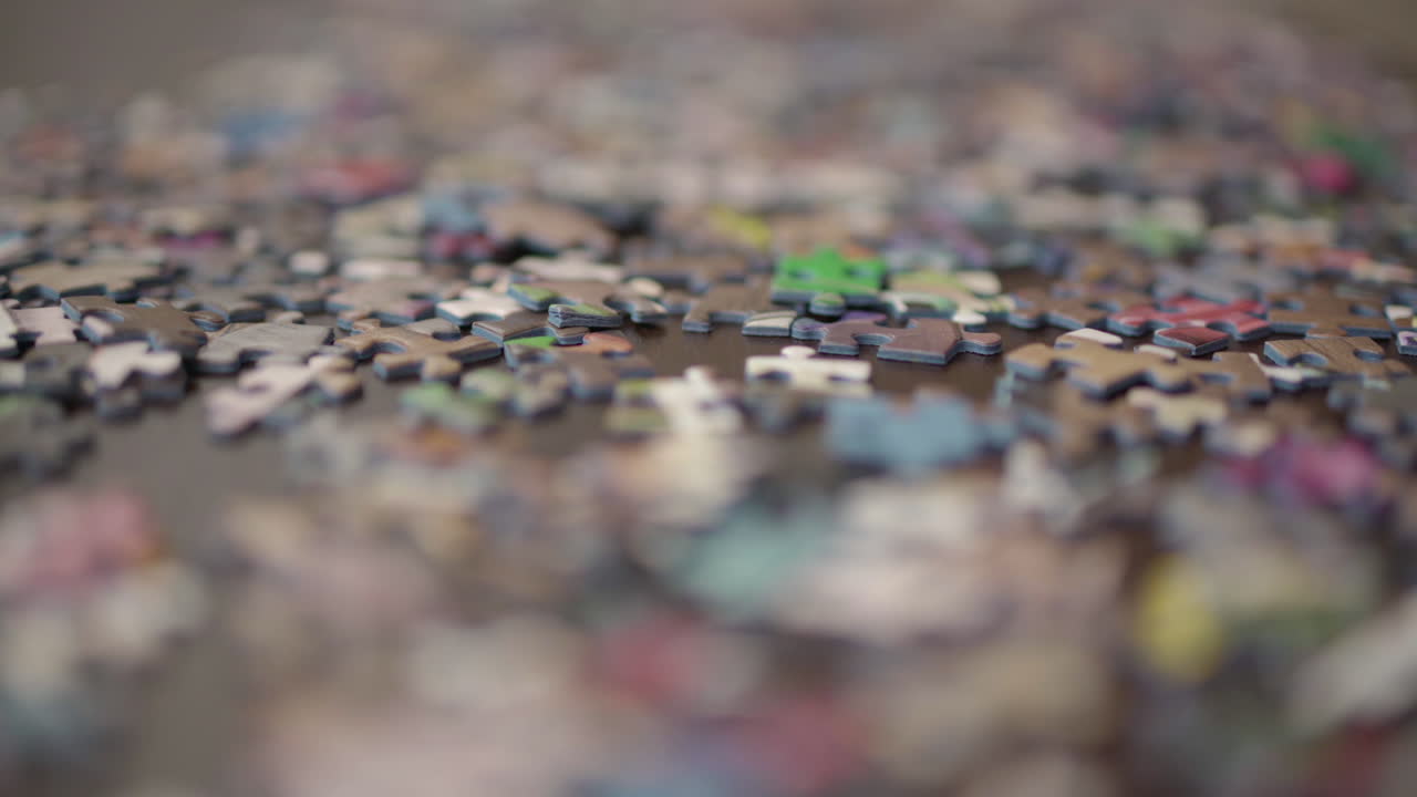 얕은 피사계 심도를 사용하여 카메라 돌리가 테이블 위에 흩어져 있는 많은 직소 퍼즐 조각을 닫습니다.