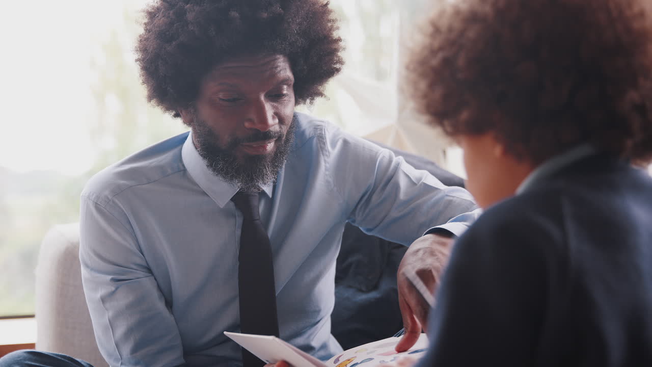 Close up of middle aged black father and his son sitting opposite each other on the sofa in their living room doing homework, selective focus
