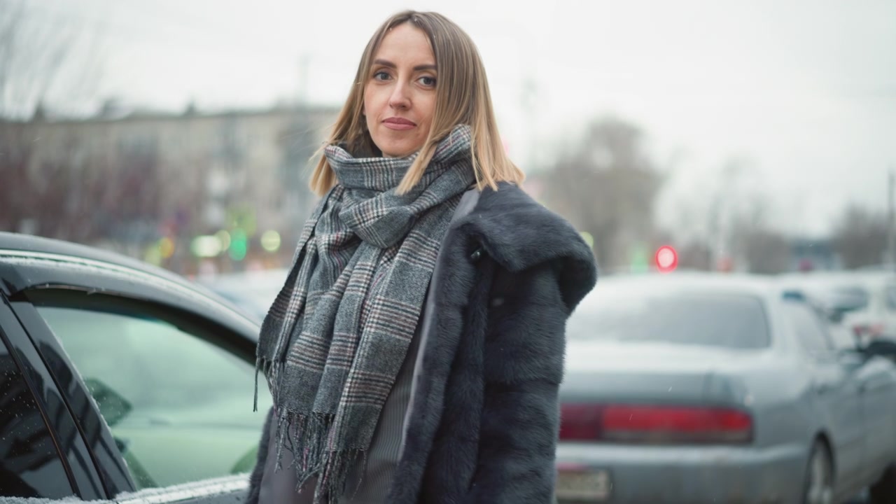 Primo piano di una donna che indossa un cappotto di pelliccia scuro e una sciarpa a quadri, in piedi vicino ad auto parcheggiate in un parcheggio esterno. Il soggetto rimane per lo più immobile, con lievi movimenti delle mani, circondata da altri veicoli.