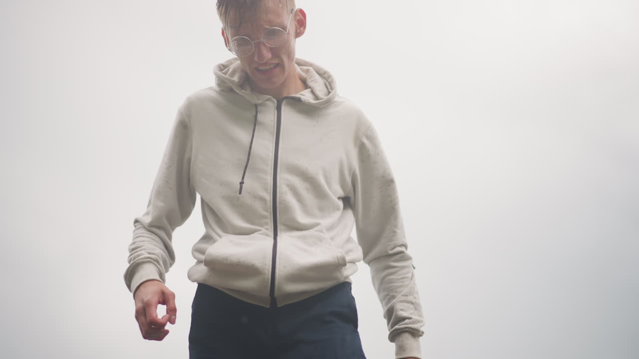 Retrato de un hombre blanco con sudadera bajo las nubes, zancada segura y mirada introspectiva junto a la orilla en un día lluvioso, joven con sudadera caminando cerca de la barandilla, pelo mojado y gotitas, pavimento reflectante