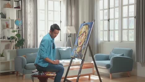 asian artist man coming to sit on round wooden chair without backrest and holding paintbrush mixed colour before painting a girl's hair on the canvas
